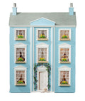 A miniature three-story dollhouse house with light blue walls, a gray roof, white trim, and tiny windows with green plants, centered on a plain white background.