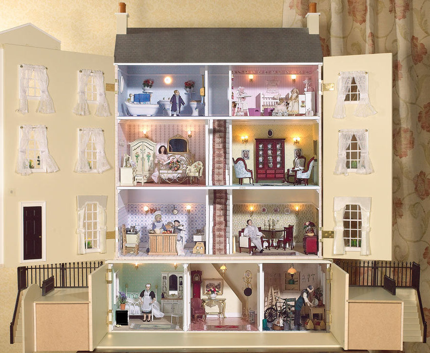 A miniature dollhouse  shown open to reveal multiple furnished rooms with tiny furniture pieces, dolls, and warm string lights. The dollhouse is displayed indoors on a wooden table against a patterned curtain background.