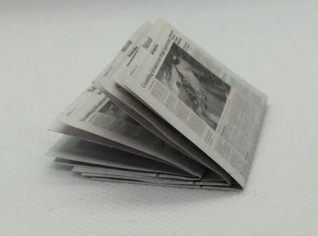 A miniature folded newspaper model, likely for dollhouse or scale modeling, placed on a white surface .