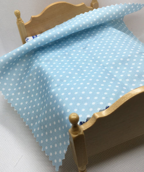 A miniature dollhouse bed with a light wood frame, styled with a light blue fabric sheet featuring a white polka dot pattern.