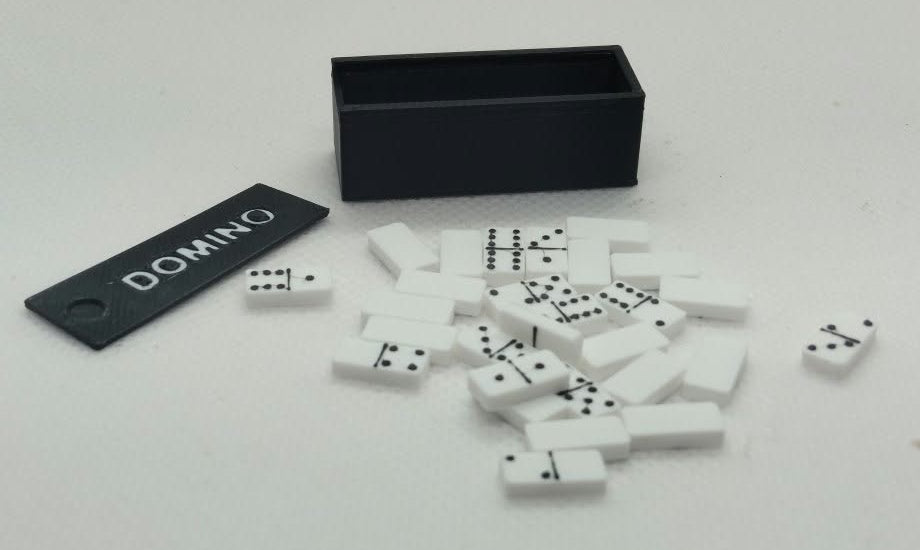 Dolls house miniature White dominoes with black pips scattered on a white surface next to a black domino box and a black tag labeled DOMINO. 