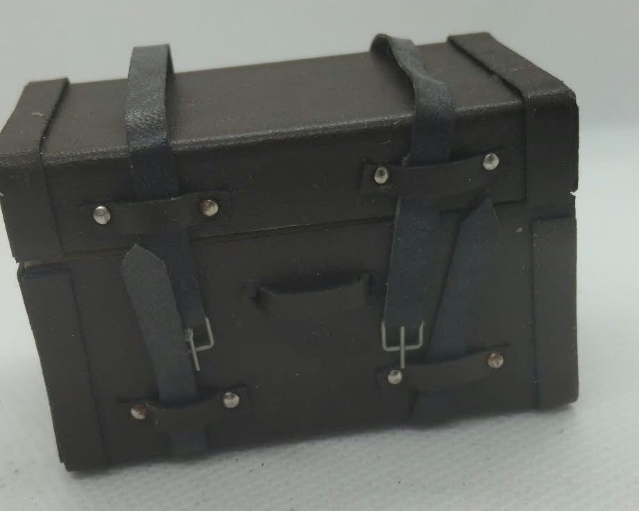 A miniature black toy chest with faux-leather straps and silver-tone rivets, photographed on a light gray background.