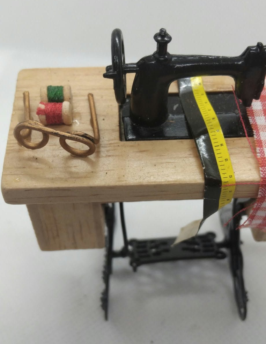 A miniature sewing machine set on a small wooden table, featuring a black sewing machine body, a yellow measuring tape, brown glasses, and miniature spools of thread, photographed against a plain light background.