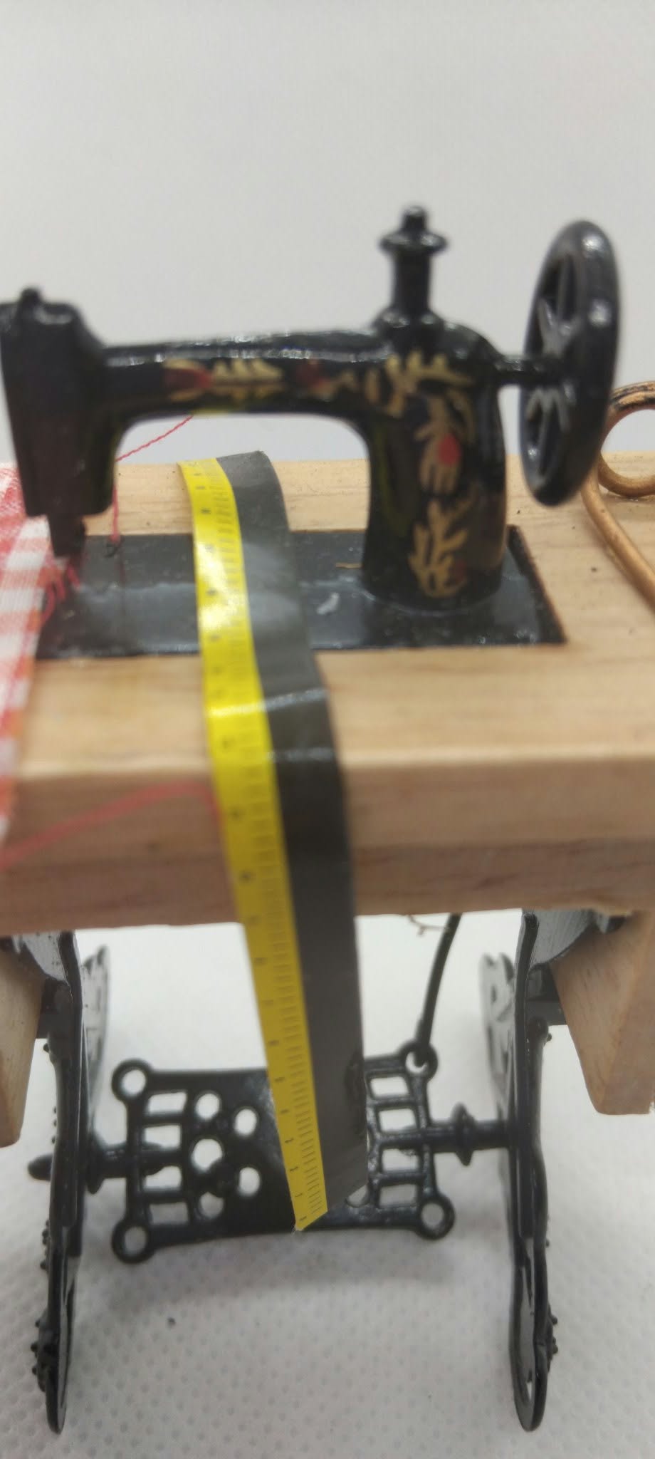 A miniature black vintage sewing machine model with gold floral accents, mounted on a light wood base with small wheels, shown with a yellow tape measure draped across it.