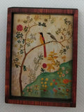 A small framed decorative painting or print showing a bird perched on a flowering tree, with a red-brown wooden frame, photographed against a plain light background.