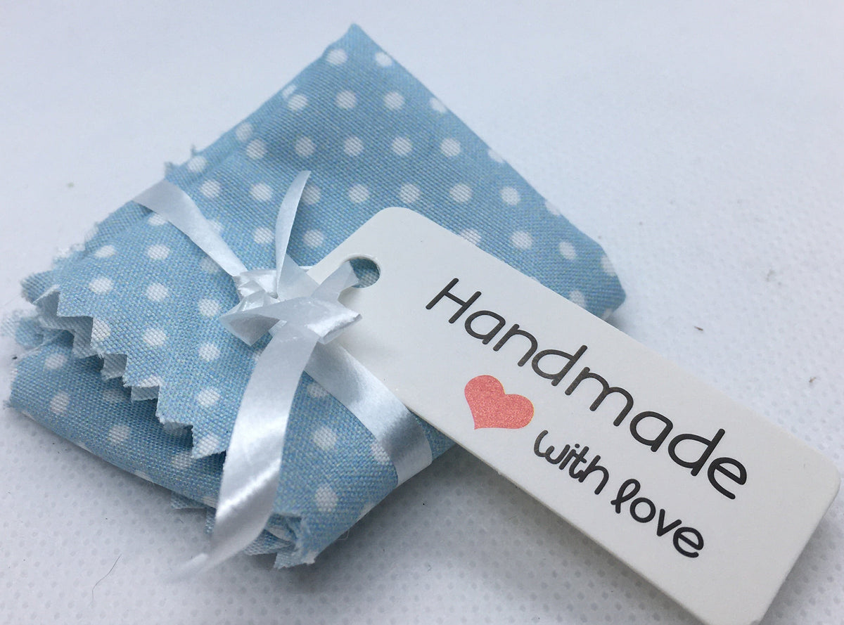 A folded light blue bed sheet in miniature with white polka dots, tied with a white ribbon and attached to a white tag that reads 'Handmade with love' with a small red heart, on a white textured background.