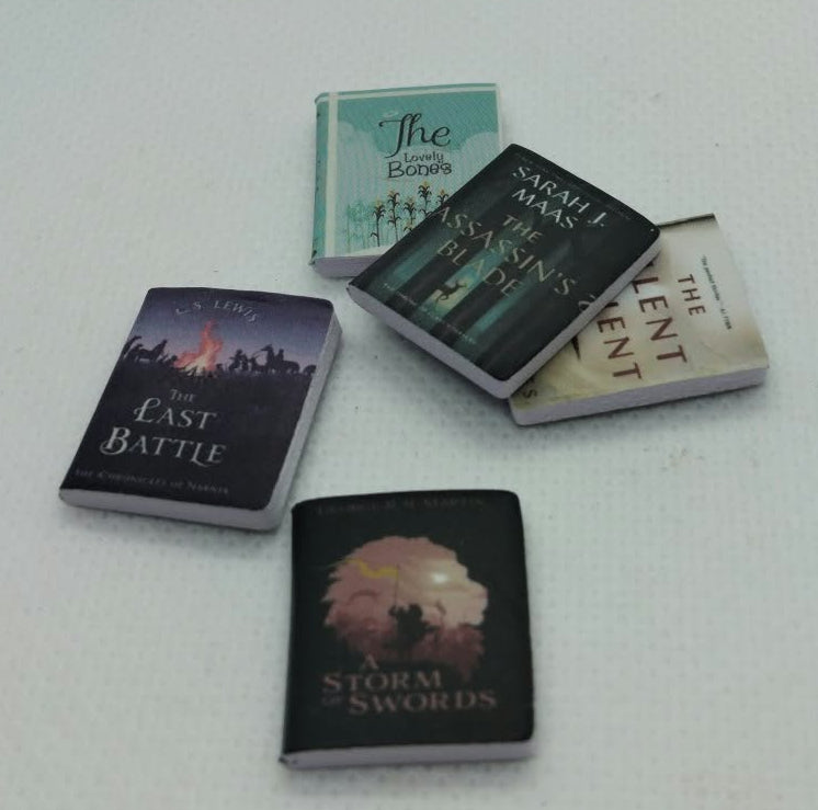 Five miniature books arranged on a plain white surface. The books have different covers, including one with a dark purple and orange sunset design, one with a black and white tree design, and one with a light green cover.
