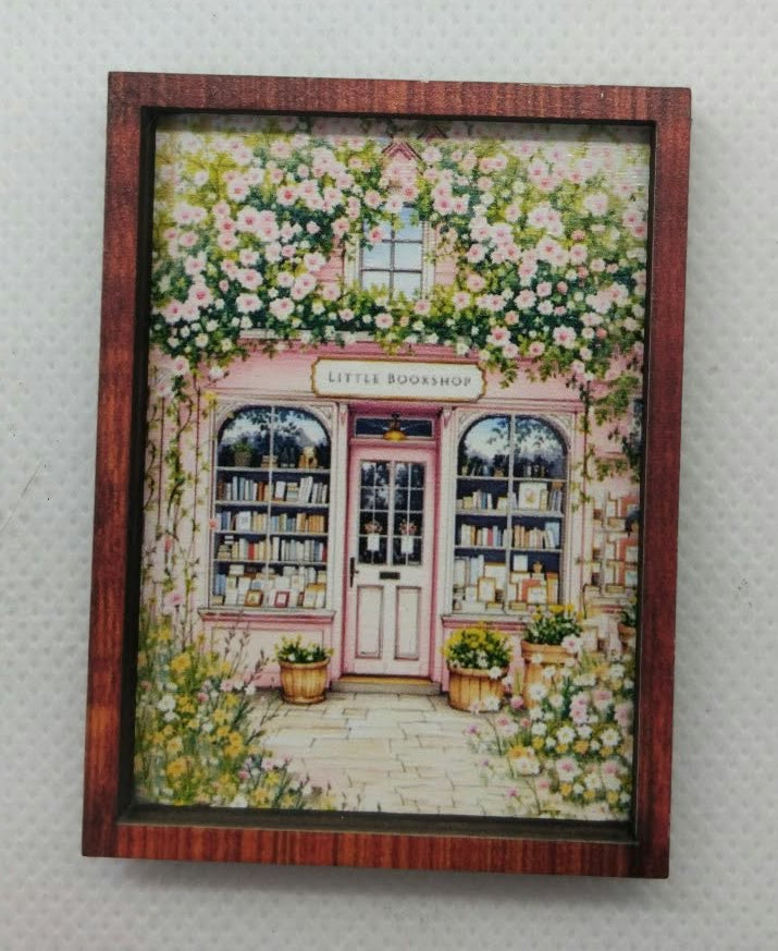 A small framed decorative print or painting showing a pink storefront with green ivy and flowers, mounted on a light textured wall.