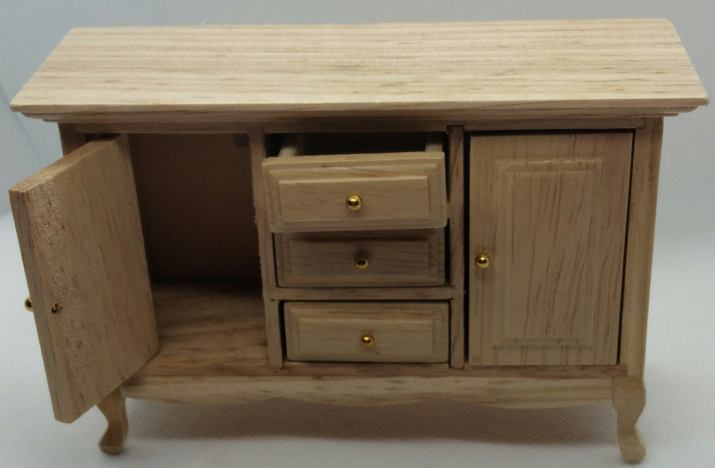 A miniature wooden cabinet or sideboard with three drawers and one open door, shown on a plain light gray background. The cabinet is made of light natural wood with small gold-colored knobs.