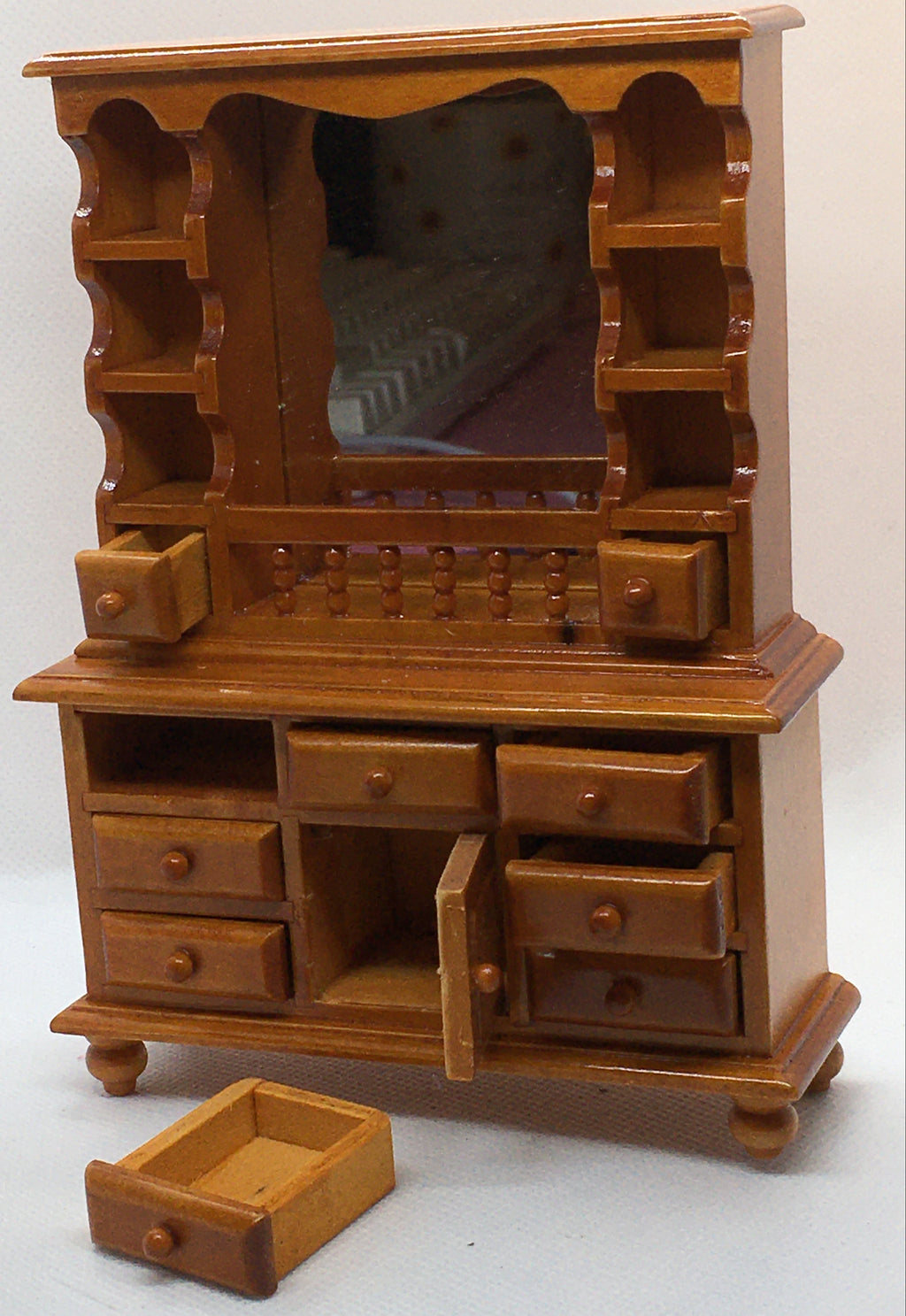 A miniature wooden vanity cabinet with an attached mirror, multiple drawers, open shelving, and one drawer pulled out in front, photographed against a plain light background.