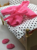 A miniature dollhouse bed with a light wood frame, white fabric bedding with black polka dots, a matching pillow, and a bright pink plush robe draped across the bed, with two small pink slippers on the floor.