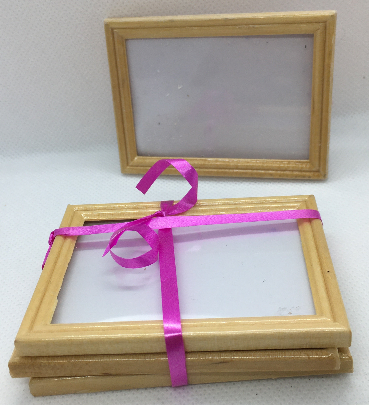 A stack of small square wooden picture frames  and a bright pink ribbon tied around the bundle, with one frame standing upright behind on a light fabric background.