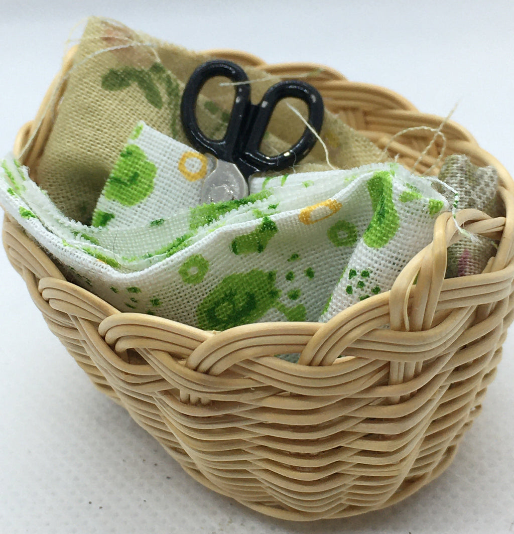 A miniature woven wicker basket containing a small beige fabric pouch with green and yellow patterns, a white fabric piece with green and yellow motifs, and black-handled scissors, set against a plain white background.