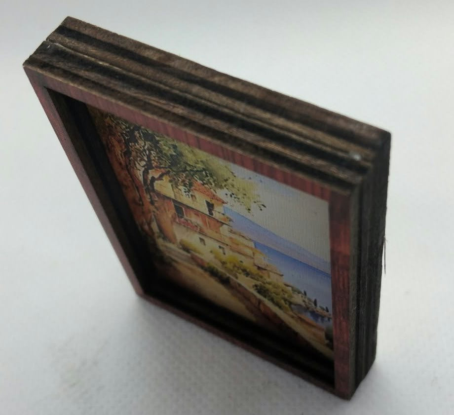 A small square framed landscape art piece with a dark brown wooden frame, standing on a light gray textured surface.
