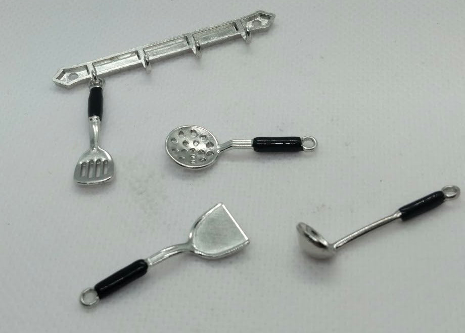 Four miniature silver-colored kitchen utensils with black handles, including a whisk, spoon, spatula, and ladle, arranged on a light gray textured surface.