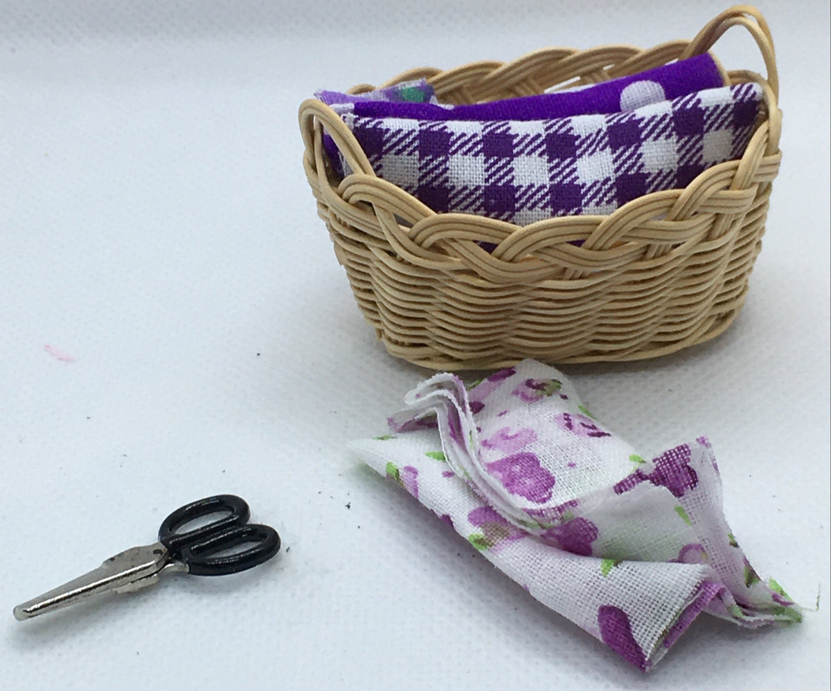 A miniature dollhouse sewing basket with a purple and white gingham fabric accompanied by a pair of tiny black-handled scissors and a small folded piece of purple floral fabric, all arranged on a white surface.