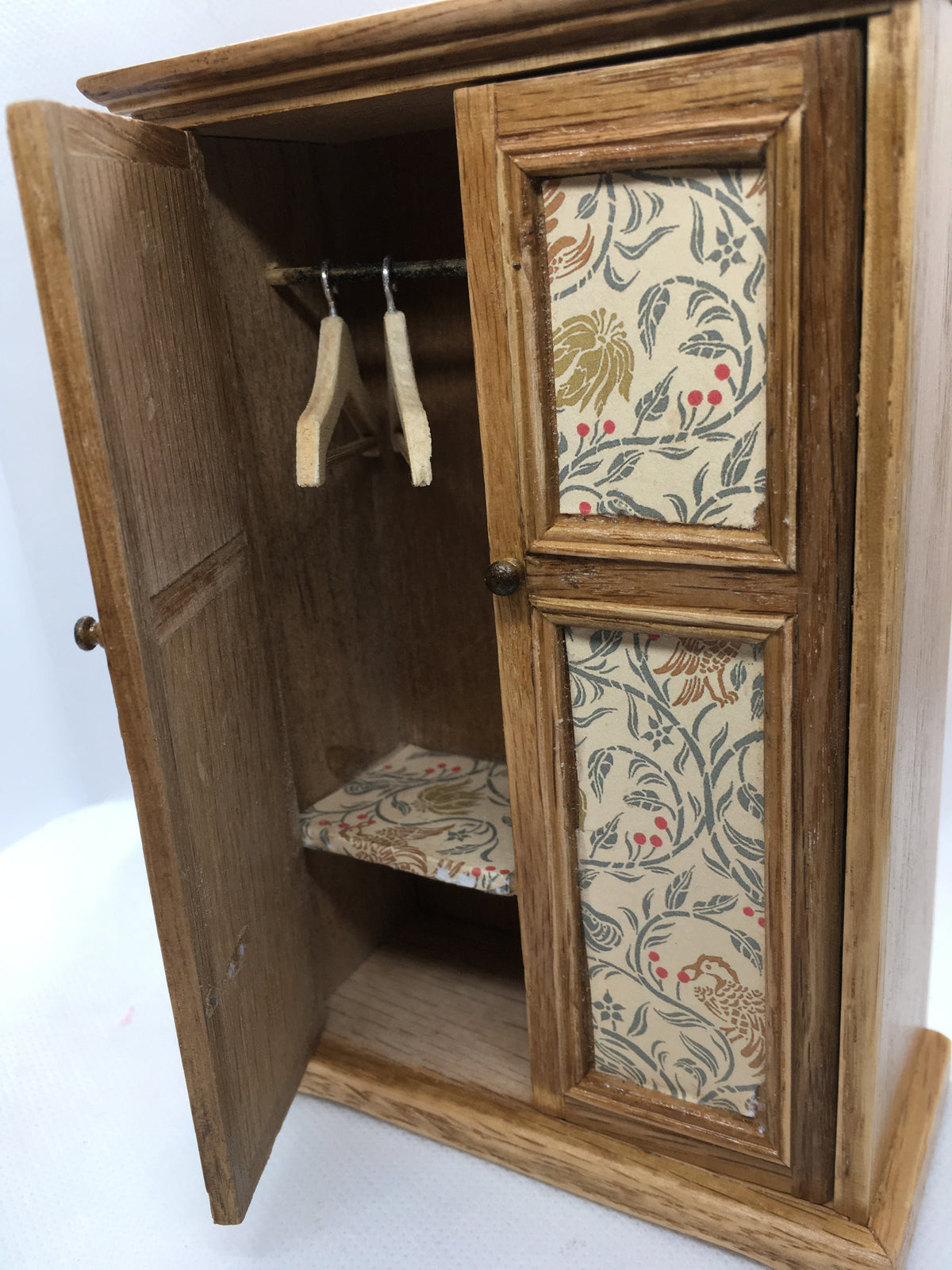 one door open on the miniature wardrobe with two coathangers and a shelf in the same vintage wallpapered material.