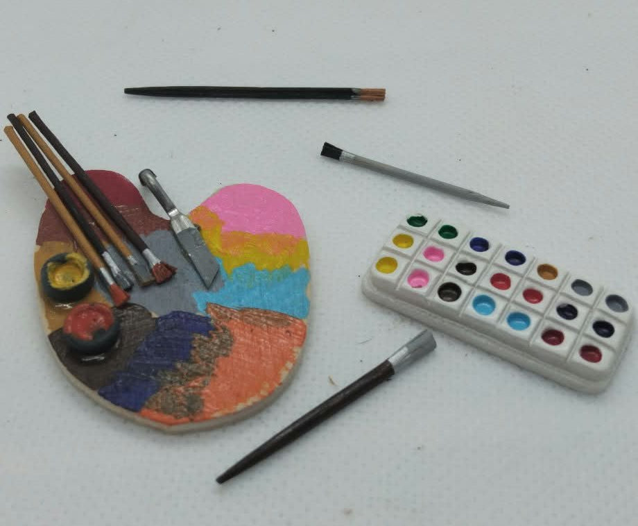 A miniature toy artist's set on a white textured background, featuring a heart-shaped paint palette with paintbrushes, a small watercolor palette, and two paint sticks.