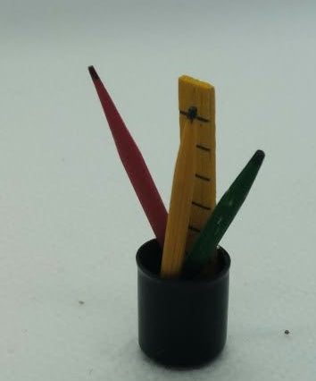 A miniature black cylindrical holder containing three small colored pencils, set on a white textured surface with a white backdrop.