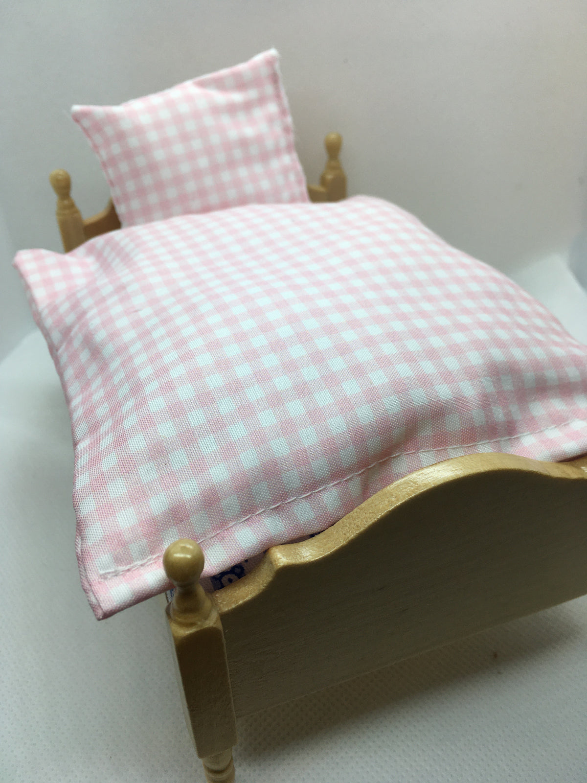 A close-up of a pink and white gingham checkered pillowcase with a matching pillow, displayed on a miniature wooden bed frame.