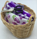 A miniature woven wicker basket holding a folded fabric with a purple and white floral pattern and a small black-handled scissors, set against a plain white background.