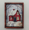 A small framed winter scene print hanging on a light gray textured wall. The artwork shows a red house with snow-covered roof and branches, a pale sky, and a snowy ground.
