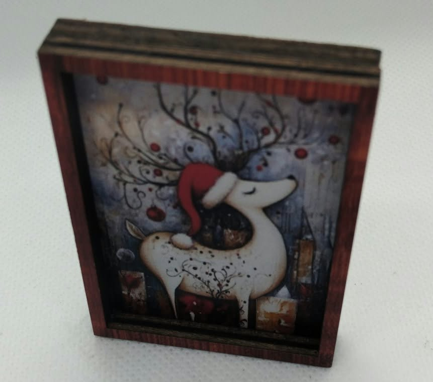 A small rectangular wooden picture frame with a Christmas-themed illustration of a white deer wearing a red Santa hat, set against a snowy forest background, photographed on a light gray surface.
